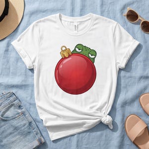 May include: A white short-sleeve t-shirt featuring a graphic of a green frog peeking out from behind a large, round, shiny red Christmas ornament. The frog has a grumpy expression with narrowed eyes, and the ornament has a gold cap.