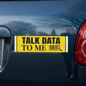 Op de afbeelding: Een rechthoekige gele bumpersticker met een zwarte rand en zwarte tekst die TALK DATA TO ME leest. Het ontwerp bevat een zwarte streepjescode naast een vergrootglas-icoon, geplaatst op de achterkant van een donkerblauw voertuig.