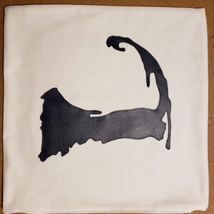 Könnte beinhalten: Cremefarbenes quadratisches Kissen mit einem schwarzen, lederartigen Applikation der Halbinsel Cape Cod. Die Applikation ist mittig platziert und hat eine detaillierte Umrissform.