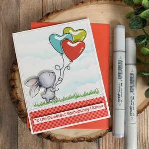 May include: A handmade greeting card featuring a gray bunny holding heart-shaped balloons in teal, red, and yellow. The card has the text "To the Sweetest Somebunny I Know" and is accompanied by Copic Sketch markers.