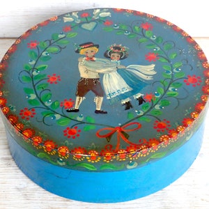 Puede incluir: Una caja redonda de madera pintada de azul con tapa. La tapa presenta una escena pintada de una pareja bailando en una corona de hojas verdes y flores rojas. La pareja lleva ropa tradicional y la mujer lleva una corona de flores en la cabeza.