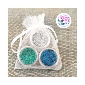 May include: Three small jars of glitter in a white organza bag. The jars contain silver, teal, and blue glitter. The bag has a white ribbon tie. The label on the bag says "Eco Glitter".
