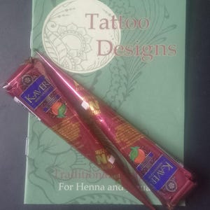 Pode incluir: Dois cones de henna Kaveri Mehendi na cor bordô colocados na diagonal sobre um livreto verde claro intitulado Tattoo Designs. Os cones apresentam etiquetas azuis e douradas com texto e um pequeno gráfico laranja. O livreto tem um logotipo branco circular.