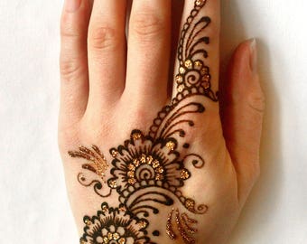 May include: A hand adorned with intricate henna designs. The dark brown patterns feature floral motifs and swirling details, accented with gold glitter. The design covers the back of the hand and extends onto the index finger.