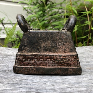 May include: A dark, weathered metal bell with two arched handles. The bell has a rectangular shape with a decorative band of etched patterns near the base. The bell is likely an antique or vintage item.