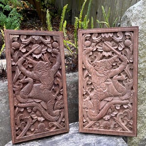 May include: Two rectangular, wooden wall panels with intricate carvings of a figure wielding a weapon, set against a floral background. The panels are a warm brown color, showcasing detailed craftsmanship.