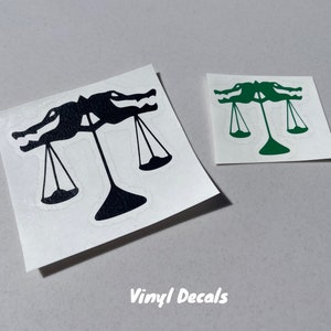 Puede incluir: Dos calcoman&iacute;as de vinilo con un dise&ntilde;o de escamas de caim&aacute;n, una negra y otra verde, que representan una balanza con dos caimanes a cada lado de la balanza.
