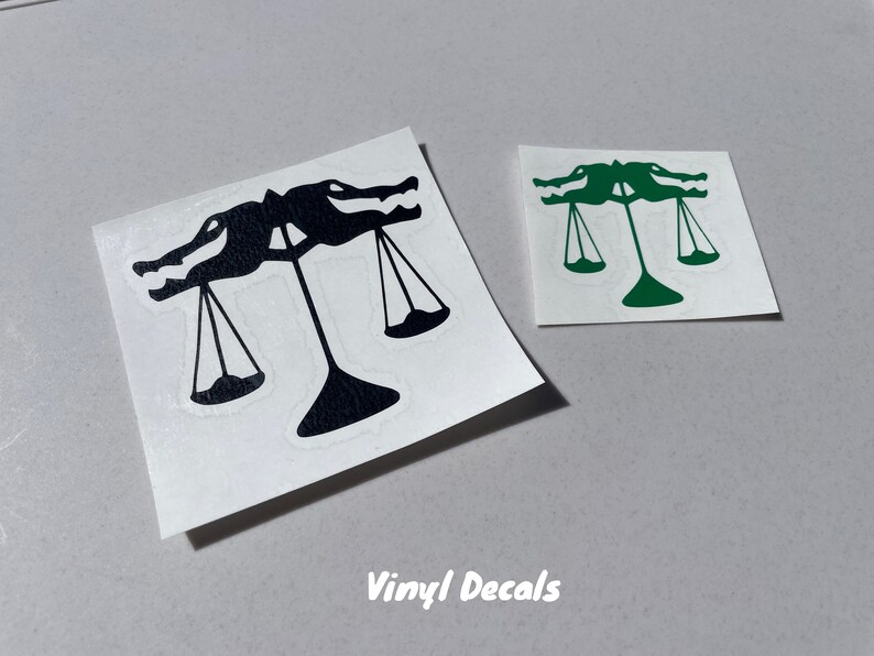 Puede incluir: Dos calcoman&iacute;as de vinilo con un dise&ntilde;o de escamas de caim&aacute;n, una negra y otra verde, que representan una balanza con dos caimanes a cada lado de la balanza.