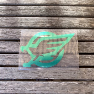 May include: A green and teal leaf-shaped sticker on a clear backing. The sticker is placed on a weathered, gray wooden surface. The design features a stylized leaf with a gradient effect, transitioning from teal to green.