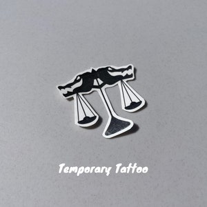 Puede incluir: Tatuaje temporal en blanco y negro de una balanza con dos cabezas de drag&oacute;n en la parte superior. El texto "Temporary Tattoo" est&aacute; impreso debajo de la imagen.