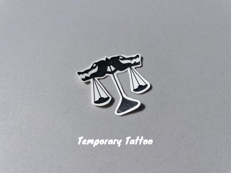 Puede incluir: Tatuaje temporal en blanco y negro de una balanza con dos cabezas de drag&oacute;n en la parte superior. El texto "Temporary Tattoo" est&aacute; impreso debajo de la imagen.