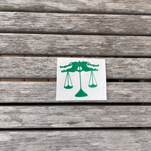 Puede incluir: Una pegatina verde con dise&ntilde;o de escamas de cocodrilo en una superficie de madera. La pegatina representa dos cocodrilos enfrentados, con sus cabezas formando la parte superior de una balanza. La balanza tiene dos pesos colgantes.