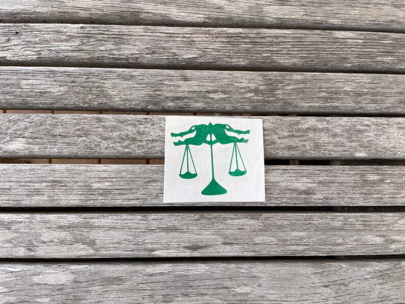 Puede incluir: Una pegatina verde con dise&ntilde;o de escamas de cocodrilo en una superficie de madera. La pegatina representa dos cocodrilos enfrentados, con sus cabezas formando la parte superior de una balanza. La balanza tiene dos pesos colgantes.