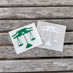 Puede incluir: Dos pegatinas de vinilo de un caim&aacute;n sosteniendo una balanza de la justicia. Una pegatina es verde y la otra es blanca.