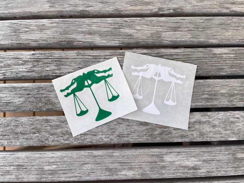 Puede incluir: Dos pegatinas de vinilo de un caim&aacute;n sosteniendo una balanza de la justicia. Una pegatina es verde y la otra es blanca.