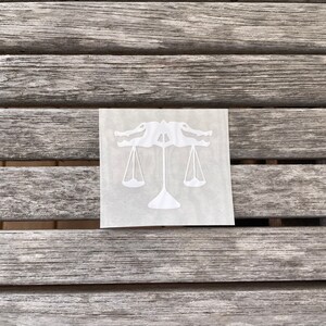 Puede incluir: Pegatina de vinilo blanco de un caim&aacute;n sosteniendo una balanza de la justicia. La pegatina est&aacute; sobre una superficie de madera.