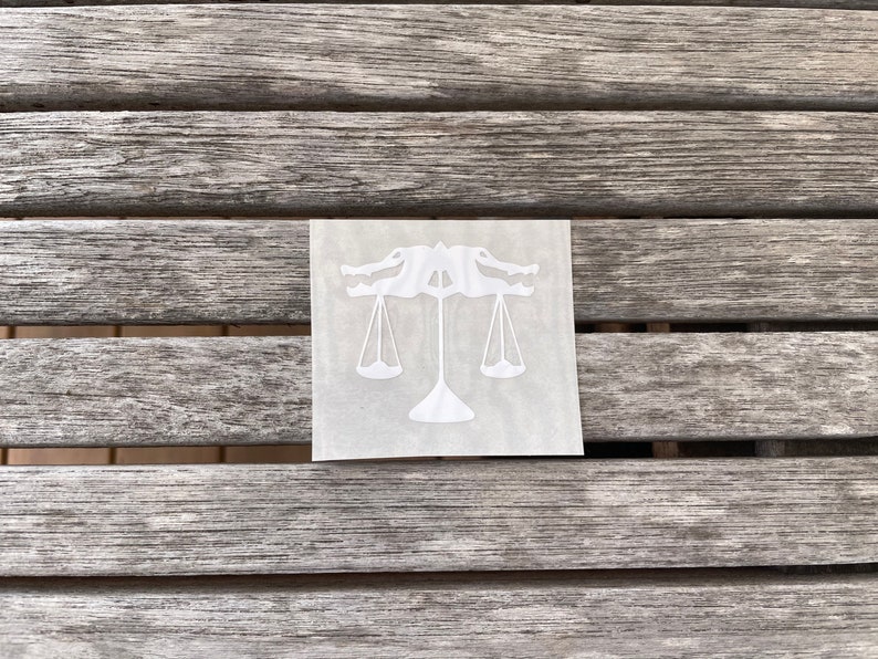 Puede incluir: Pegatina de vinilo blanco de un caim&aacute;n sosteniendo una balanza de la justicia. La pegatina est&aacute; sobre una superficie de madera.