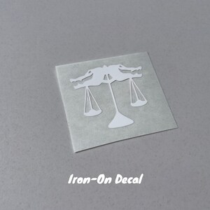 Puede incluir: Calcoman&iacute;a blanca para planchar de una balanza con dos figuras sosteniendo las balanzas. El texto "Iron-On Decal" est&aacute; impreso debajo de la imagen.