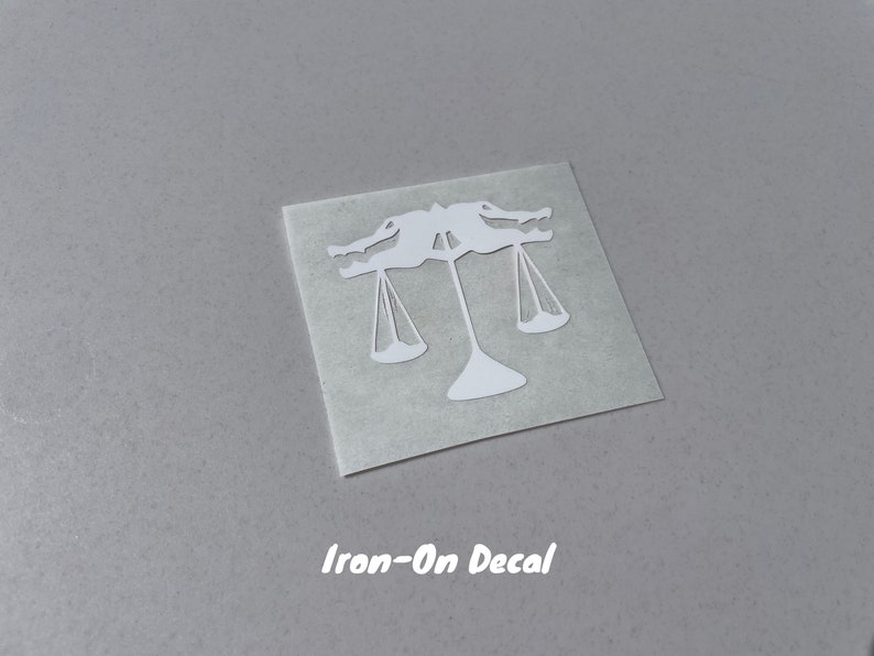 Puede incluir: Calcoman&iacute;a blanca para planchar de una balanza con dos figuras sosteniendo las balanzas. El texto "Iron-On Decal" est&aacute; impreso debajo de la imagen.