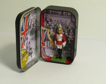 Könnte beinhalten: Geöffnete kleine schwarze Dose mit einer Miniaturfigur eines britischen Soldaten. Das Innere der Dose zeigt Bilder der Flagge Union Jack und eine Menschenmenge. Der Soldat trägt eine rote Jacke, weiße Hose und einen goldenen Helm.