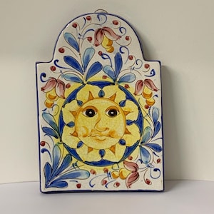 May include: A rectangular ceramic wall plaque with a rounded top, featuring a hand-painted yellow sun face at the centre surrounded by blue and red floral motifs on a white background with a visible crackle glaze finish and a blue border.