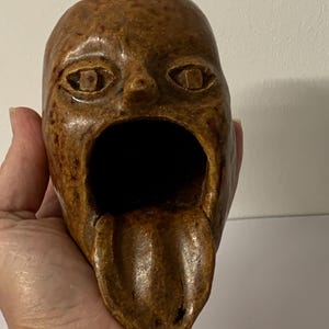 May include: A brown, speckled ceramic sculpture shaped like a human head with a wide, open mouth and a protruding tongue. The surface has a glossy, textured finish with visible dark brown spots throughout the clay form, featuring carved eyes.