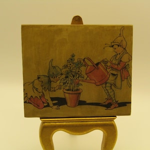 May include: A square decorative tile featuring a vintage-style illustration of two elves tending to a potted plant. One elf kneels while the other stands holding a red watering can. The tile is displayed on a gold-coloured stand with curved legs.