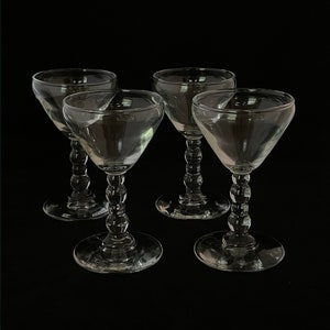 May include: Four clear glass stemmed cocktail glasses. Each glass has a round base, a beaded stem, and a triangular bowl. The glasses are arranged against a black background.