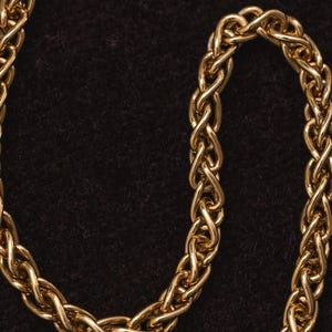 May include: A close-up of a gold chain necklace with a braided design. The chain is lying on a dark brown surface.