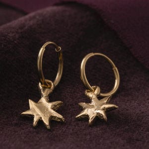 May include: A pair of gold hoop earrings with a star charm hanging from each hoop. The earrings are on a dark purple velvet surface.