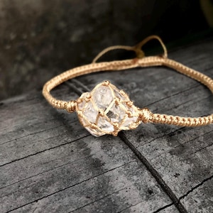 May include: A gold-colored macrame bracelet featuring a clear quartz crystal. The crystal is encased in a woven gold-toned net, and the bracelet is set against a weathered wooden surface.