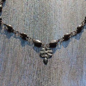 May include: A silver necklace with a pendant featuring a flower design. The necklace is made of small, oval-shaped beads connected by a chain.