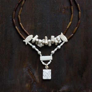 May include: A handcrafted necklace featuring a layered design. The necklace has two strands of dark brown wooden beads. The focal point includes white coral-like pieces, fish-shaped accents, and a carved rectangular pendant. The necklace is set against a dark wood background.