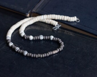 May include: A gray and white beaded necklace with a silver accent. The necklace is made of round beads and is lying on a dark surface.