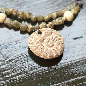May include: A necklace with a large, grey, fossilized shell pendant. The necklace is made of green and white beads.