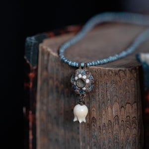 May include: A delicate necklace with a blue beaded chain and a pendant. The pendant features a floral design with white and blue accents, and a white, tulip-shaped drop. The necklace rests on a wooden surface.