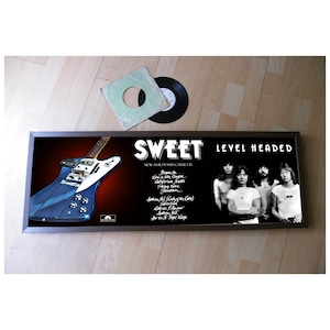 Puede incluir: Una portada de álbum enmarcada para el álbum "Level Headed" de la banda Sweet. La portada presenta una guitarra eléctrica azul y una foto en blanco y negro de los miembros de la banda.