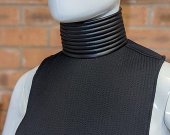 May include: A black faux leather collar with a textured fabric backing. The collar is designed to be worn around the neck and has a high, stand-up style.