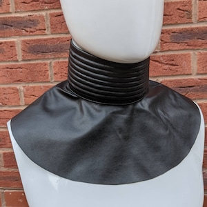 May include: A black faux leather collar with a ribbed design. The collar is worn on a white mannequin.