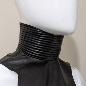 May include: A black leather collar with a ribbed design. The collar is worn on a white mannequin.