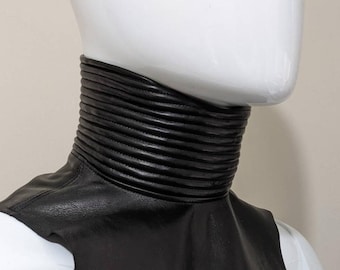 May include: A black leather collar with a ribbed design. The collar is worn on a white mannequin.