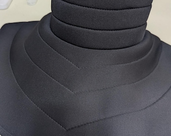 May include: Black neck brace with a layered design. The brace features a series of horizontal padded sections around the neck and a wider, contoured base. A zipper is visible at the back of the neck. The item is made of a soft, matte material.