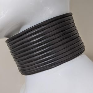 May include: A black leather choker necklace with a wide, layered design. The choker is made of multiple horizontal bands, each with a stitched detail. The necklace is displayed on a white mannequin.