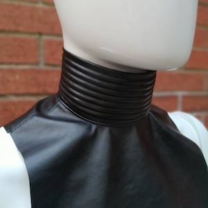 May include: A black faux leather collar with a ribbed design. The collar is worn on a white mannequin.