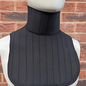 May include: A black neck brace with vertical stitching, displayed on a white mannequin head. The brace has a high collar and a bib-like front, with a rounded bottom edge. The background is a brick wall.