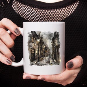 May include: White ceramic mug featuring a watercolor illustration of a cobblestone street with buildings and streetlights. The artwork depicts a European town scene with a moody, atmospheric quality. The mug is held by a person.