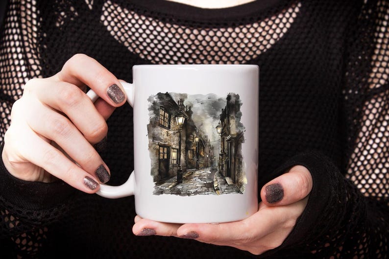 May include: White ceramic mug featuring a watercolor illustration of a cobblestone street with buildings and streetlights. The artwork depicts a European town scene with a moody, atmospheric quality. The mug is held by a person.