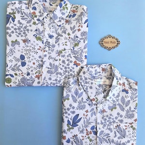 Peut inclure: Deux chemises blanches à motifs floraux bleus, verts et oranges. Les chemises sont pliées sur une surface bleu clair. Une petite étiquette en bois avec l'inscription "Hand Made" est également présente.