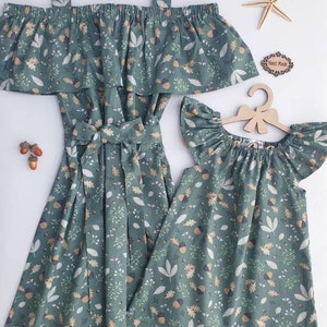 May include: Matching green dresses with a leaf and acorn pattern. One dress is off-the-shoulder with straps, and the other is a smaller, flutter-sleeved dress. A starfish and a wooden sign that says "Hand Made" are also in the image.