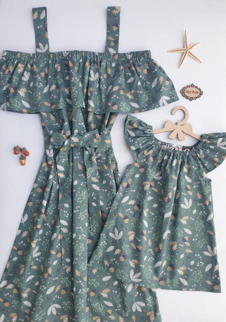 May include: Matching green dresses with a leaf and acorn pattern. One dress is off-the-shoulder with straps, and the other is a smaller, flutter-sleeved dress. A starfish and a wooden sign that says "Hand Made" are also in the image.